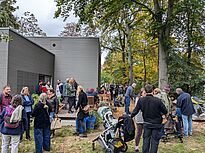 Vor dem Gebäude und zwischen hohen Bäumen haben sich viele Erwachsene und Kinder versammelt. Einige Menschen stehen auf einer Holzterrasse, andere sitzen an Tischen oder schieben Kinderwagen über den mit Holzschnitzeln bedeckten Außenbereich.