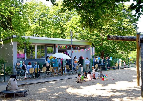 Vor dem Frauen-Café sitzen Personen gemeinsam an Tischen, Kinder spielen im Sand.