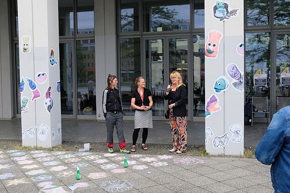 Drei Personen richten bei der Ausstellungseröffnung Grußworte an die Anwesenden, an den Säulen neben ihnen hängen Bilder von den MINIMAs.