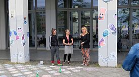 Drei Personen richten bei der Ausstellungseröffnung Grußworte an die Anwesenden, an den Säulen neben ihnen hängen Bilder von den MINIMAs.