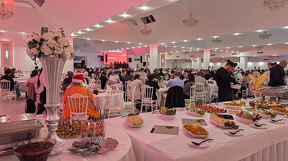 In einem festlich beleuchteten Saal sitzen zahlreiche Gäste an gedeckten Tischen; vorne am Bildrand stehen Blumen und ein Buffet mit verschiedenen Speisen.