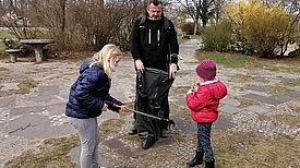 Ein Mann und zwei Kinder sammeln auf einer Freifläche Müll; ein Kind nutzt eine Greifzange.