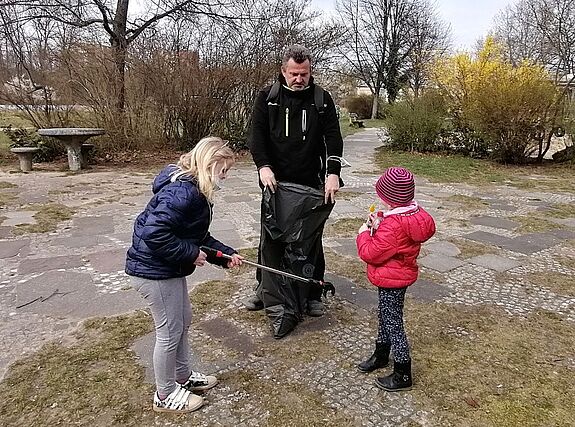 Ein Mann und zwei Kinder sammeln auf einer Freifläche Müll; ein Kind nutzt eine Greifzange.
