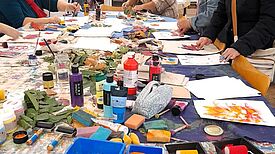 Mehrere Personen sitzen an einem großen Tisch und bedrucken Stoffbeutel mit buntem Herbstlaub. Auf dem Tisch liegen Farben, Pinsel, Walzen und bemalte Blätter.