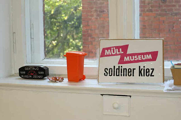 Ein Fensterbrett mit einem kleinen orangefarbenen Mülleimer und einem Schild mit der Aufschrift „Müll Museum Soldiner Kiez“.