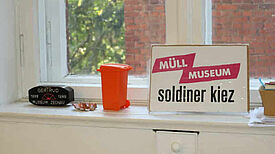 Ein Fensterbrett mit einem kleinen orangefarbenen Mülleimer und einem Schild mit der Aufschrift „Müll Museum Soldiner Kiez“.