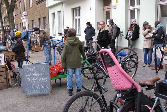 Menschen stehen mit Fahrrädern vor einem Gebäude, während ein Schild auf einen kostenlosen Fahrrad-Check hinweist.