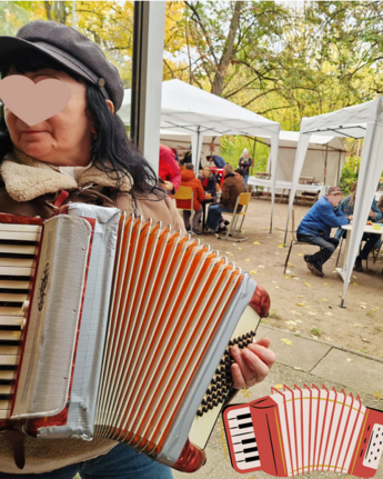 Eine Frau spielt Akkordeon im Freien, im Hintergrund sitzen Menschen unter Zelten bei einem Fest.