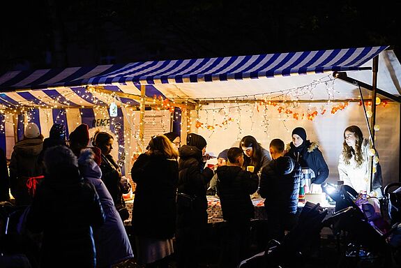 Einige Menschen stehen vor einem erleuchteten Marktstand auf dem Lichtermarkt im QM-Gebiet Klixstraße / Auguste-Viktoria-Allee.