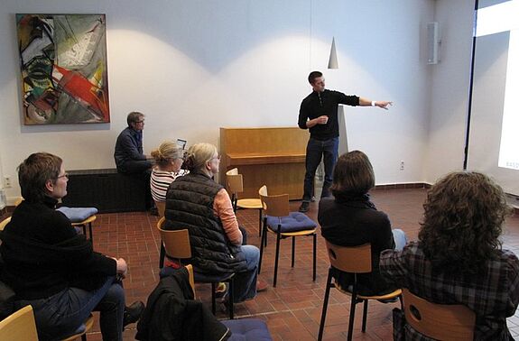 Eine Person zeigt in einem Saal auf eine Präsentation, mehrere Personen sitzen davor auf Stühlen.