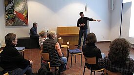 Eine Person zeigt in einem Saal auf eine Präsentation, mehrere Personen sitzen davor auf Stühlen.