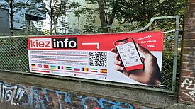 Ein Banner der „kiezinfo“-App mit QR-Code und mehrsprachiger Beschreibung hängt an einem Zaun im QM-Gebiet Zentrum Kreuzberg.
