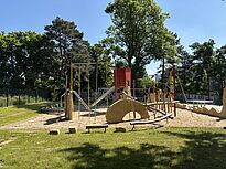 Im Vordergrund liegt ein großer Spielbereich mit Sandboden, Kletterstangen, Balancierbalken, einer Rutsche und einem roten Spielturm. Zwischen den Geräten bewegt sich ein Kind, während hohe Bäume und ein Zaun den Hintergrund bilden.