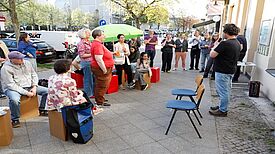 Auf einem Gehweg hören Menschen im Stehen oder Sitzen einem Mann zu, der durch ein Mikrophon spricht.  