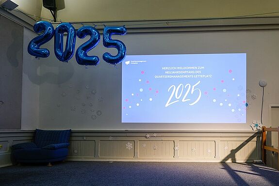 Auf der Bühne hängen Zahlen-Luftballons des aktuellen Jahres: „2025“, zudem ist eine Präsentation zum Neujahresempfang an die Wand projiziert. 