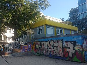 Eine Metalltreppe führt zu einem einstöckigen Containerbau mit mehreren Fenstern und einem gelben Dach. Die Box steht auf einer Mauer, die mit Graffiti besprüht ist. Im Hintergrund Wohnbebauung und ein großer Baum.