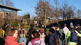 Eine Gruppe von ca. zwanzig Erwachsenen von hinten sichtbar auf einer begrünten Freifläche. Zu ihnen spricht eine Person von vorn sichtbar. Im Hintergrund Bäume und ein moderner Bau, an dem der Schriftzug „Wohntheke“ angebracht ist.   