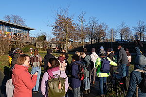 Eine Gruppe von ca. zwanzig Erwachsenen von hinten sichtbar auf einer begrünten Freifläche. Zu ihnen spricht eine Person von vorn sichtbar. Im Hintergrund Bäume und ein moderner Bau, an dem der Schriftzug „Wohntheke“ angebracht ist.   