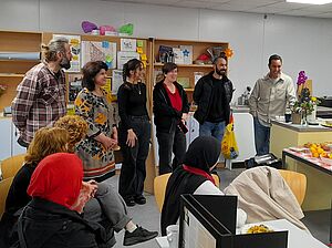 In einem Raum stehen und sitzen mehrere lachende Personen vor einem Tisch, auf dem Lebensmittel stehen. 