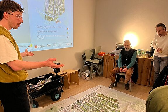 Ein Mann spricht zu anderen Menschen, die um einen großen, auf dem Boden liegenden Quartiersplan sitzen und stehen. Ein Quartiersplan ist an die Wand projiziert. 