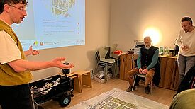 Ein Mann spricht zu anderen Menschen, die um einen großen, auf dem Boden liegenden Quartiersplan sitzen und stehen. Ein Quartiersplan ist an die Wand projiziert. 