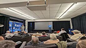 Menschen sitzen auf Sofas vor einer Leinwand, auf der zwei Personen die Entwürfe für das neue Stadtteilzentrum präsentieren.
