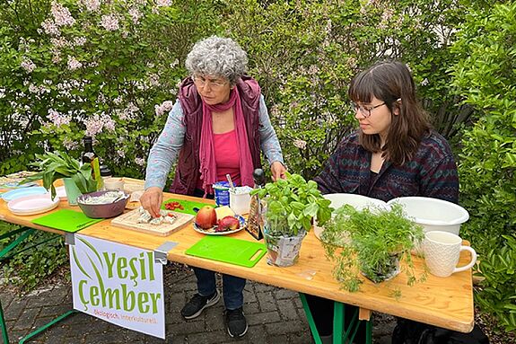 Zwei Personen bereiten an einem Tisch im Freien frische Brotaufstriche mit Kräutern und Obst zu, vorne hängt ein Schild mit der Aufschrift „Ye?il Çember“.