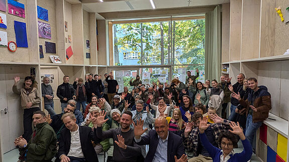 Viele Menschen sitzen und stehen in einem hellen Raum und winken gemeinsam in die Kamera.