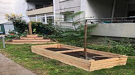 Zwei Schaufeln liegen auf einem mit Erde befüllten Hochbeet vor einem Wohnhaus. Daneben steht ein Pyramidenbeet.