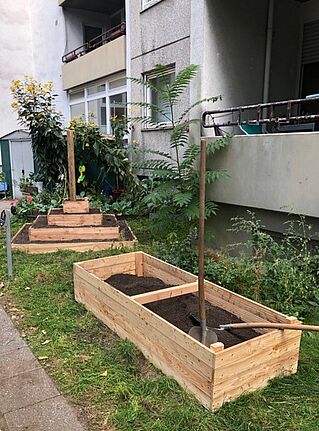Zwei Schaufeln liegen auf einem mit Erde befüllten Hochbeet vor einem Wohnhaus. Daneben steht ein Pyramidenbeet.