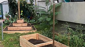 Zwei Schaufeln liegen auf einem mit Erde befüllten Hochbeet vor einem Wohnhaus. Daneben steht ein Pyramidenbeet.