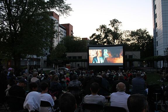 Menschen schauen bei Einbruch der Dunkelheit in einem Wohnviertel gemeinsam einen Film auf einer großen Leinwand.