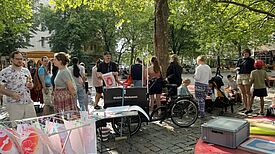 Menschen stehen an einer mobilen Siebdruckwerkstatt auf einem Platz unter Bäumen neben einem Wäscheständer, auf dem bedruckte Blätter und T-Shirts trocknen, und Tischen mit Arbeitsmaterialien.