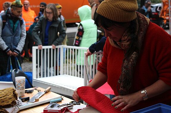 Künstlerin in rotem Pullover repariert beim BSR-Kieztag im Harzer Kiez ein Möbelstück, umgeben von Besucherinnen und Besuchern.