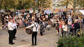 Musiker spielen auf Trommeln und Blasinstrumenten, während Kinder und Erwachsene unter Girlanden aus bunten Bändern feiern.