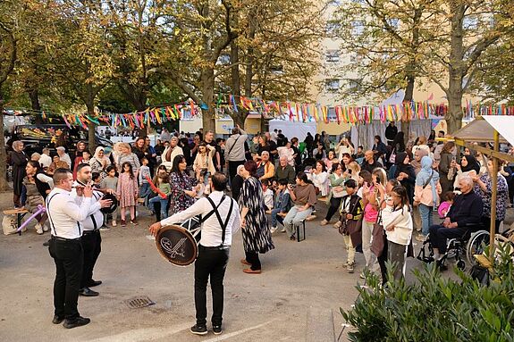 Musiker spielen auf Trommeln und Blasinstrumenten, während Kinder und Erwachsene unter Girlanden aus bunten Bändern feiern.