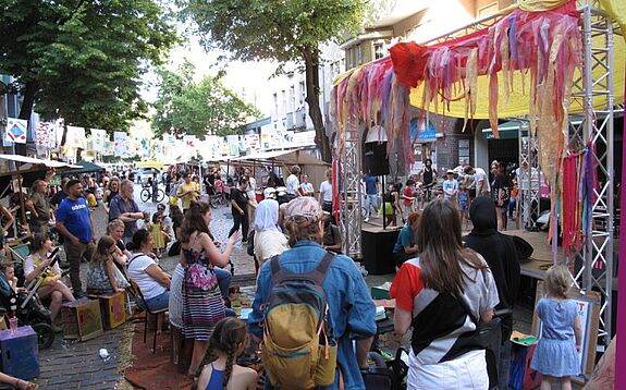 Viele Menschen stehen und sitzen vor einer Bühne beim Kiezfest in der Weichselstraße im QM-Gebiet Donaustraße-Nord.