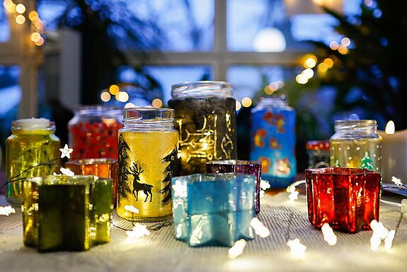 Bunte, selbst gestaltete Windlichter aus Glas stehen auf einem Tisch und leuchten inmitten von Lichterketten.