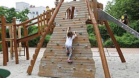 Auf einem Spielplatz klettert ein Kind eine Schrägwand aus Holz hoch, die zu einer Rutsche und einer Holzbrücke führt, auf der weitere Kinder sind. Die Kindergesichter sind mit Sternen anonymisiert. 