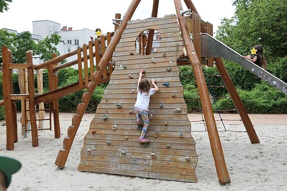 Auf einem Spielplatz klettert ein Kind eine Schrägwand aus Holz hoch, die zu einer Rutsche und einer Holzbrücke führt, auf der weitere Kinder sind. Die Kindergesichter sind mit Sternen anonymisiert. 