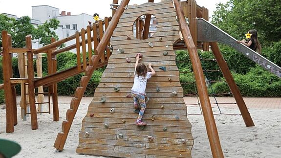 Auf einem Spielplatz klettert ein Kind eine Schrägwand aus Holz hoch, die zu einer Rutsche und einer Holzbrücke führt, auf der weitere Kinder sind. Die Kindergesichter sind mit Sternen anonymisiert. 