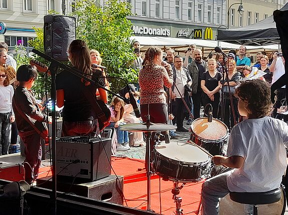 Eine Kinderband spielt auf einer Open-Air-Bühne. Rechts befindet sich der Schlagzeuger, links die Gitarristen, in der Mitte steht die Sängerin. Das Foto ist von der Rückseite der Bühne aus aufgenommen, vor der Bühne steht das Publikum. 
