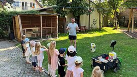 Kinder stehen in einer Reihe im Kita-Garten und hören einem Mann zu, der eine Pflanzaktion erklärt.