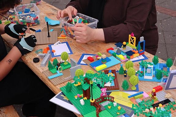 Auf einer Bierzeltgarnitur unter freiem Himmel sitzen Menschen und basteln mit LEGO-Bausteinen.