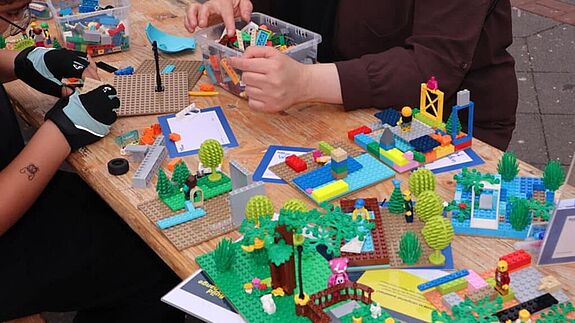 Auf einer Bierzeltgarnitur unter freiem Himmel sitzen Menschen und basteln mit LEGO-Bausteinen.