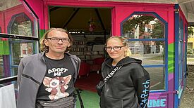 André Wien und die Koordinatorin stehen vor dem bunt gestrichenen Gemeinschaftspavillon „Zur Spinne“ am Boulevard Kastanienallee, im Hintergrund sind Bücherregale sichtbar.