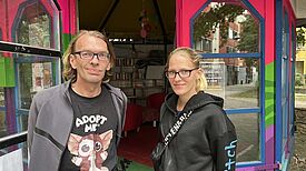André Wien und die Koordinatorin stehen vor dem bunt gestrichenen Gemeinschaftspavillon „Zur Spinne“ am Boulevard Kastanienallee, im Hintergrund sind Bücherregale sichtbar.