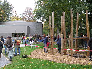 Kinder und Erwachsene an einem Klettergerät aus Holz, das mit einem rot-weißen Flatterband umgrenzt ist. Eine neue Rasenfläche und gepflasterte Wege sind sichtbar, im Hintergrund ein Funktionsneubau aus Holz und viele Menschen.