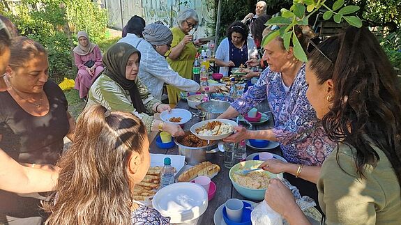 Menschen aus der Nachbarschaft treffen sich an einer langen Tafel im Gemeinschaftsgarten Prinzessinengärten und essen gemeinsam die gekochten Speisen.