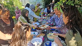 Menschen aus der Nachbarschaft treffen sich an einer langen Tafel im Gemeinschaftsgarten Prinzessinengärten und essen gemeinsam die gekochten Speisen.